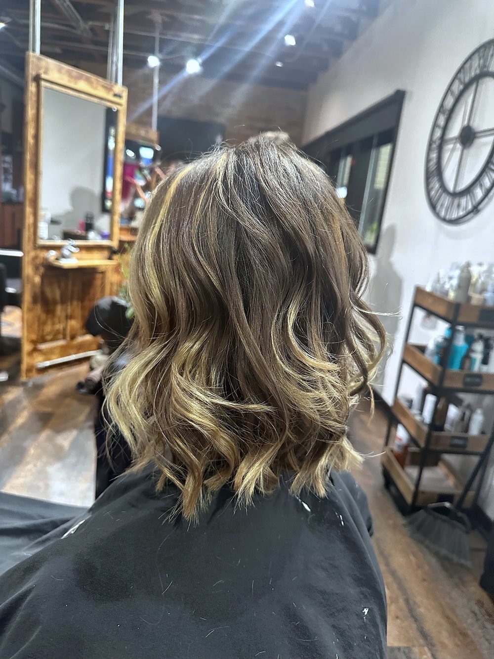 Back view of Tracie's guest at the chair: shoulder-length waves with blonde pieces through brown, the wall clock and wooden mirrors around her.